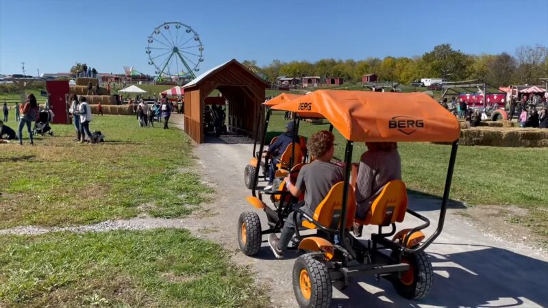 Visitors Ride Orange Pedal Carts Across a Wooden Bridge at The Fall Harvest Festival at Heaven Hill Farm, with A Ferris Wheel and Various Farm Activities Visible in The Background